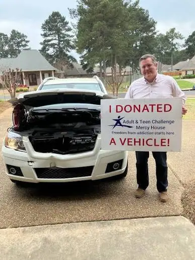 A donor beside a vehicle with the hood raised, holding an 'I donated a vehicle' sign